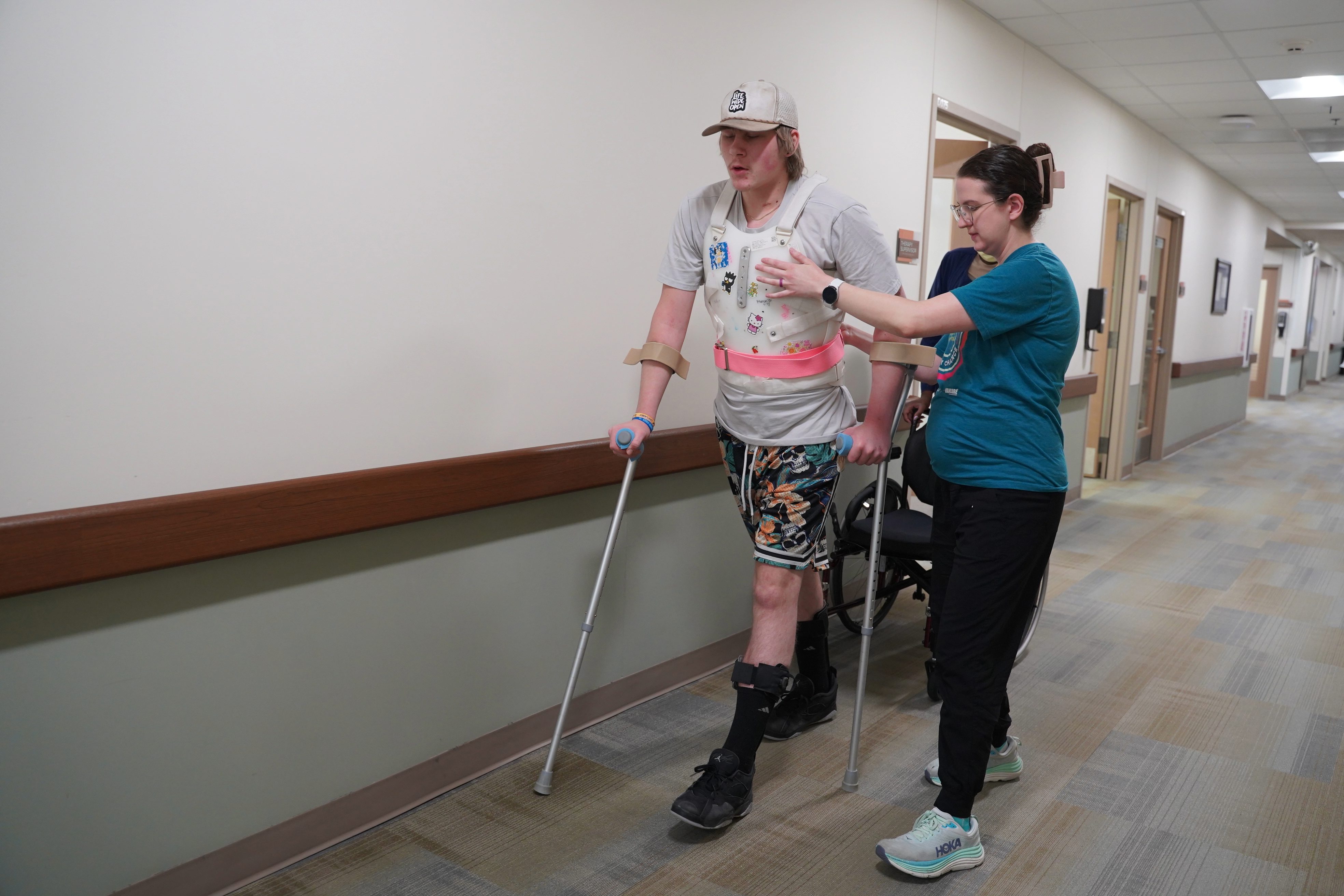 Boy using crutches walks through a hallway in a clinical setting. He is being assisted by one of Madonna's physical therapists.